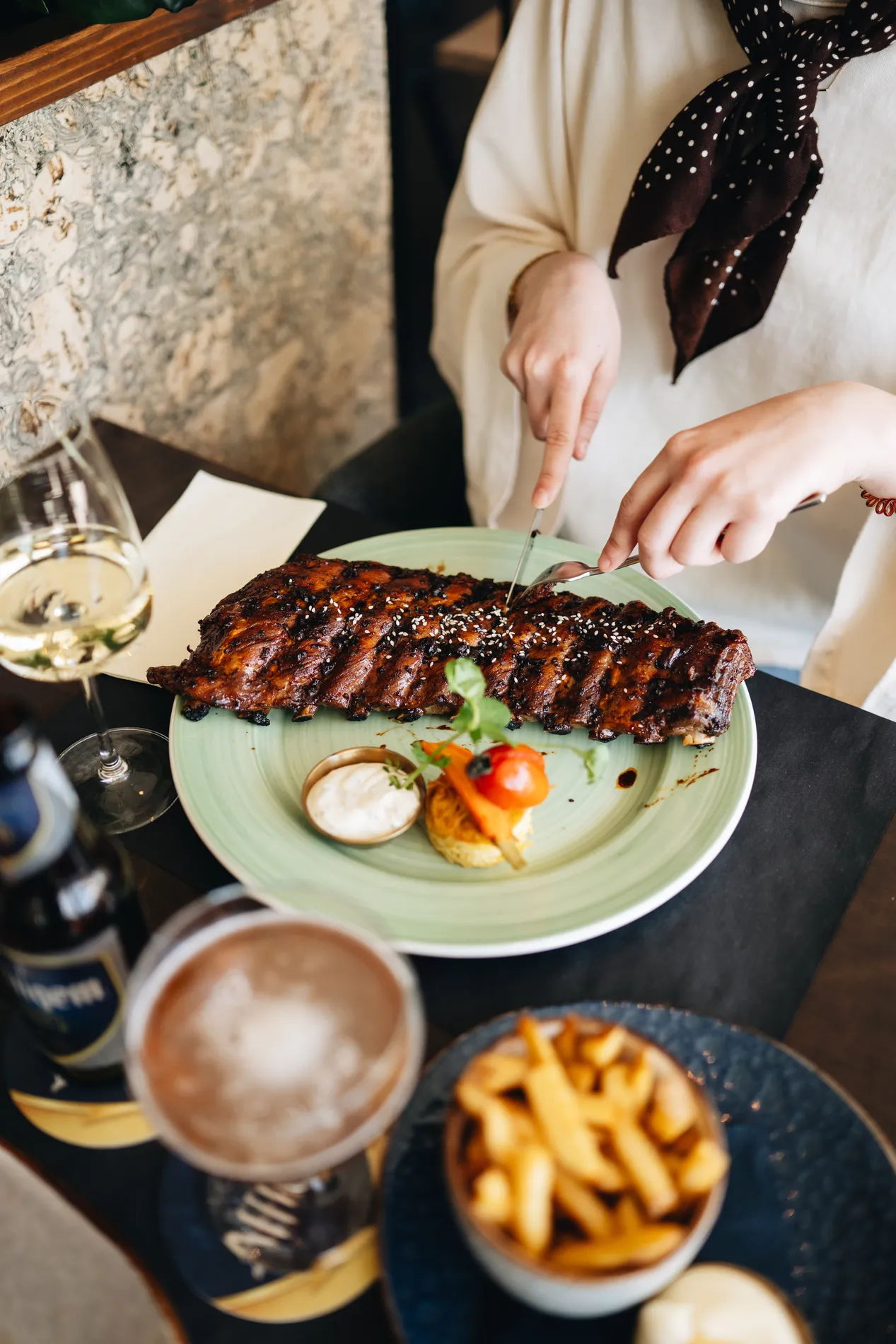 Close-up van een gast die een grote streng spareribs met sesamzaadjes aansnijdt op een lichtgroen bord, met op de voorgrond een glas witte wijn en een schaaltje frites.
